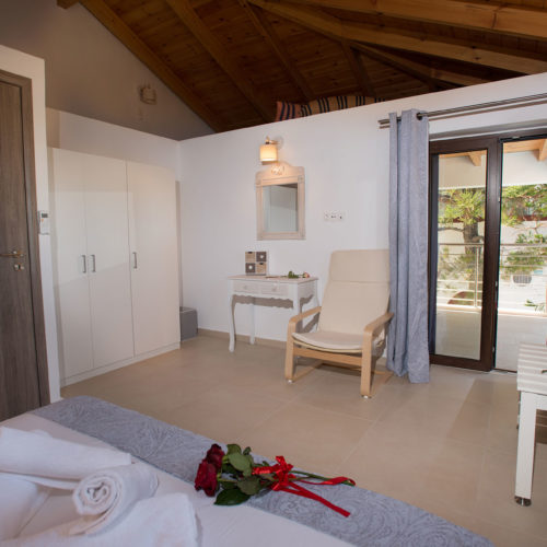 A bright bedroom with a wooden vaulted ceiling, balcony doors, a cream armchair, and neatly made bed linens