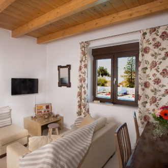 Cozy living room with wooden ceiling beams, floral curtains, a sofa, armchair, and window view at Houmeli Apartments