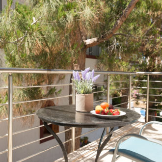 A sunny balcony with a small round table, fruit plate, potted flowers and two chairs beside pine trees