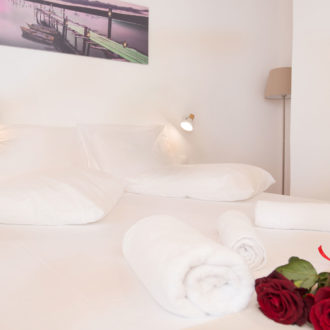 Bright white bedroom with neatly made bed, rolled towels, bedside lights, and red roses at Houmeli Apartments