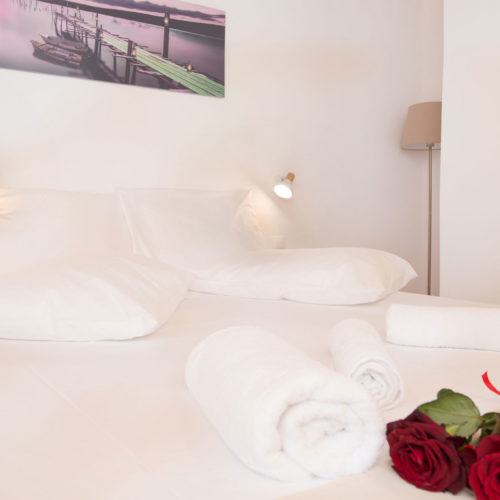 Bright white bedroom with neatly made bed, rolled towels, bedside lights, and red roses at Houmeli Apartments
