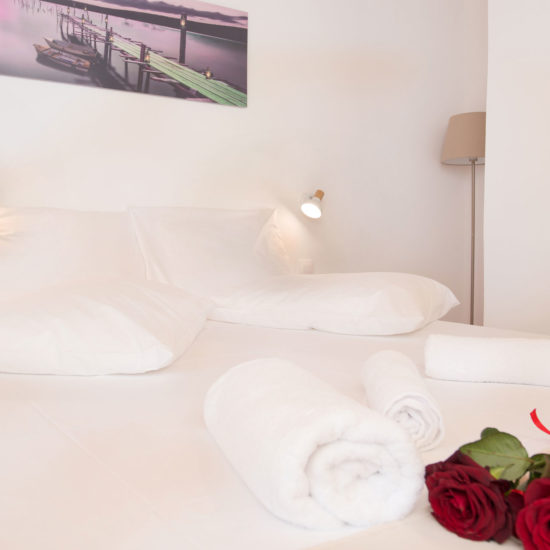 Bright white bedroom with neatly made bed, rolled towels, bedside lights, and red roses at Houmeli Apartments