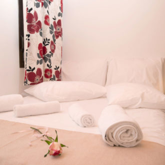 A neatly made bedroom with white bedding, rolled towels, a bedside lamp and floral curtains