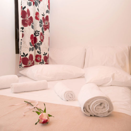 A neatly made bedroom with white bedding, rolled towels, a bedside lamp and floral curtains