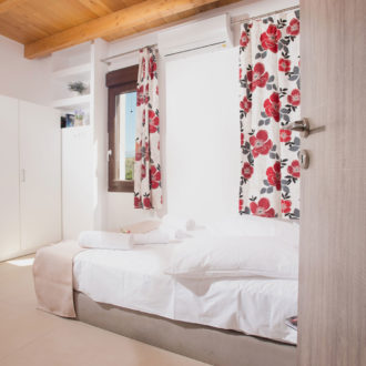 A bright ground-floor bedroom with a white bed, floral curtains, built-in wardrobes, and wooden ceiling beams