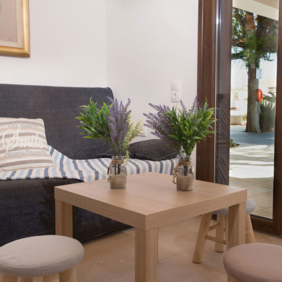 A cozy lounge corner with a sofa, small wooden table, and open glass door leading to a shaded patio