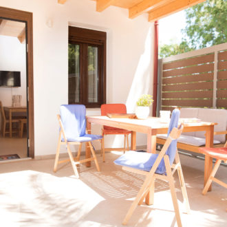 Covered patio with a wooden dining table, colorful chairs, and an open doorway into the apartment