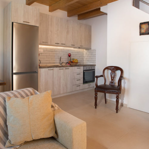 A cozy maisonette living area with a compact kitchenette, striped sofa, wooden chairs and exposed beams