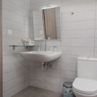 A clean modern bathroom with tiled walls, wall-mounted sink, mirror, toilet, and wooden door at Houmeli Apartments