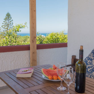 Wooden terrace table with wine, fruit, glasses and a sea view beside a cushioned corner seat