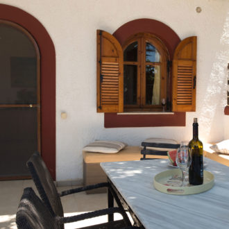 Shaded patio with a marble table, woven chairs, cushioned bench seating, and wooden shutters at Skinos apartment