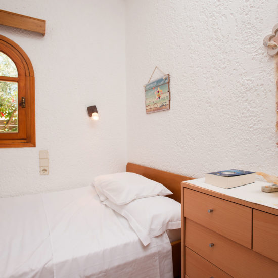 A bright single bedroom with white textured walls, a wooden dresser, and an arched window looking out to the garden