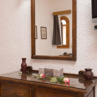 A wooden dresser with a mirror, wall light, and small decorative vases against a textured white wall