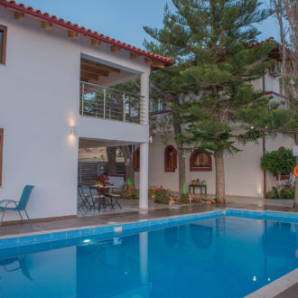 Courtyard pool beside white apartment buildings with trees, seating, and a calm evening atmosphere at Houmeli Apartments