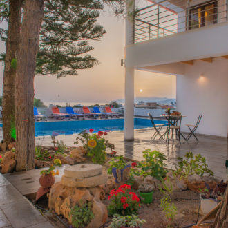Courtyard pool area with lounge chairs, trees, and a small seating table beside a white apartment building at sunset