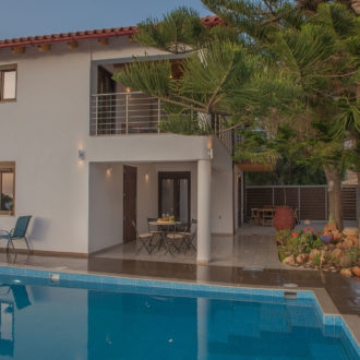 Courtyard pool beside a white two-story building with shaded seating, a tree, and patio space at Houmeli Apartments
