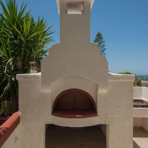 White outdoor barbecue oven in a sunny garden area with blue sky, greenery, and tiled paving at Houmeli Apartments