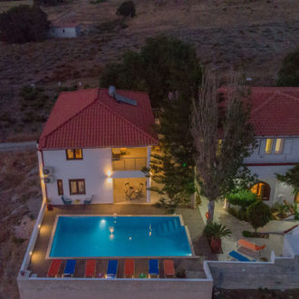 Aerial dusk view of Houmeli Apartments courtyard with a lit pool, red-roofed buildings and quiet countryside surroundings