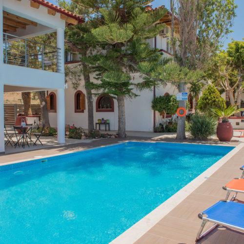 Outdoor pool area at Houmeli Apartments in Kalamaki, Crete, with sun loungers, shaded seating, and trees around the deck