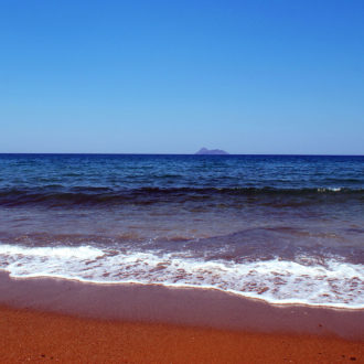 Waves on the sandy shore of Kalamaki Beach in Crete with clear blue sea and an island on the horizon.