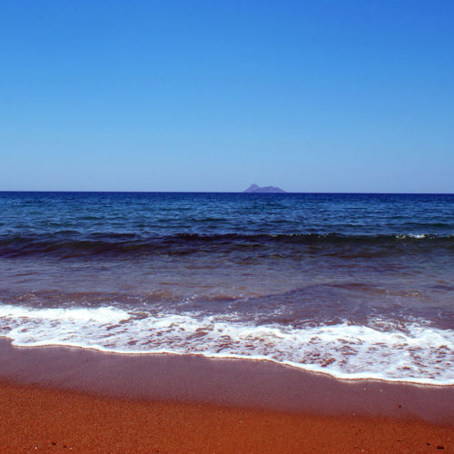 Waves on the sandy shore of Kalamaki Beach in Crete with clear blue sea and an island on the horizon.