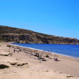 Sandy Kommos beach with sun umbrellas, swimmers, and rocky cliffs under a clear blue sky