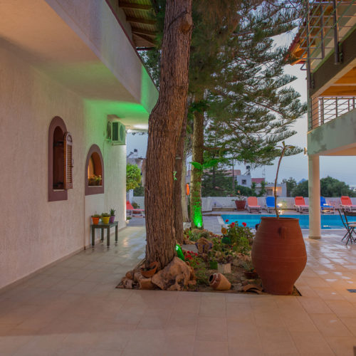 Courtyard walkway beside Houmeli Apartments with trees, potted plants and a pool area beyond