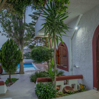 Courtyard walkway beside a pool at Skinos Apartment with trees, lounge chairs, and a lit doorway