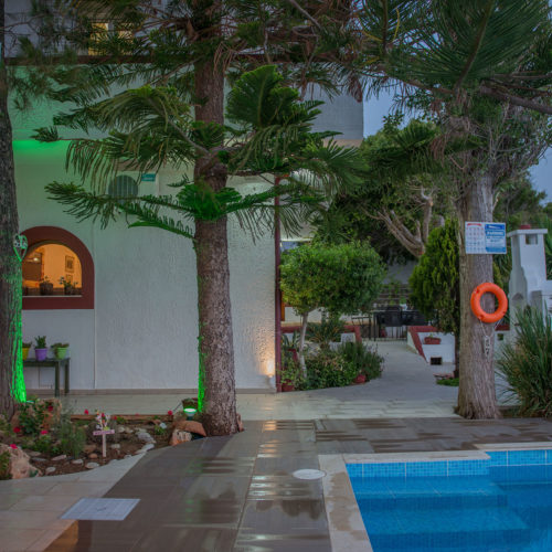 Courtyard pool area at Houmeli Apartments with trees, tiled walkway, and white buildings in Kalamaki Crete