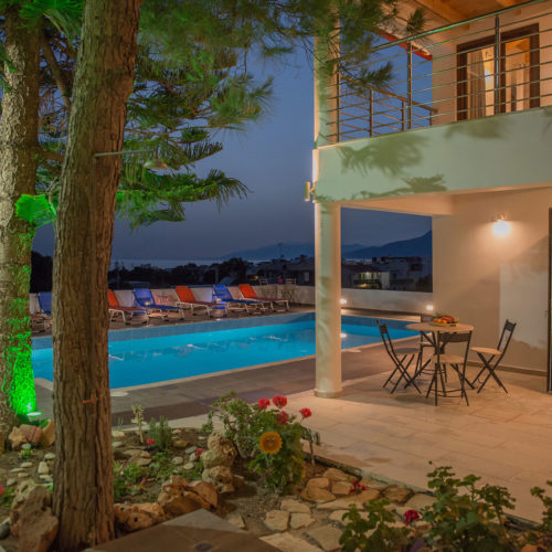 Night view of a lit apartment courtyard with a swimming pool, terrace seating, and palm trees