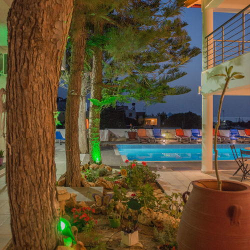 Night view of a landscaped pool area with tall trees, lounge chairs, and a balcony at Houmeli Apartments