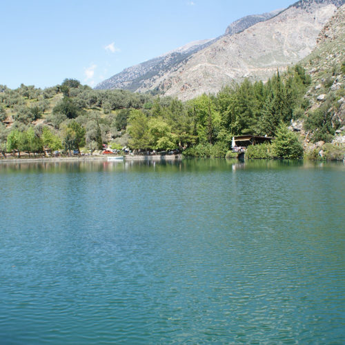 Mountain lake with calm water, trees along the shore, and rocky slopes under a clear sky