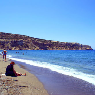 People relaxing on a sandy beach with clear blue sea and rocky cliffs under a bright sky