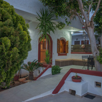 Shaded courtyard at Houmeli Apartments with white walls, trees, and outdoor seating