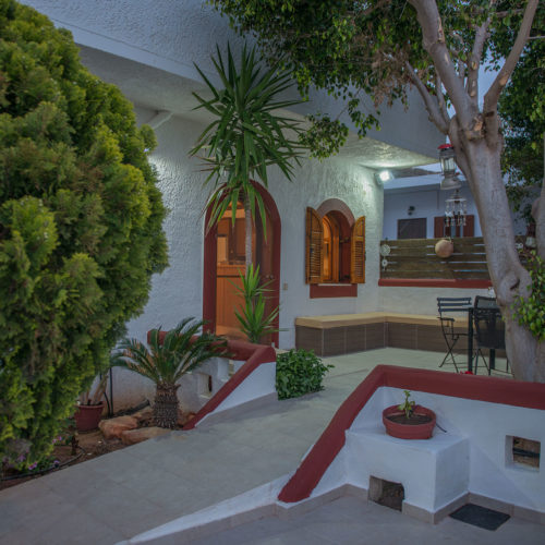 Shaded courtyard at Houmeli Apartments with white walls, trees, and outdoor seating