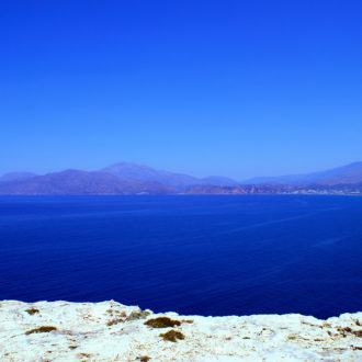 Wide view of a calm blue sea with distant mountains and a rocky shoreline in the foreground