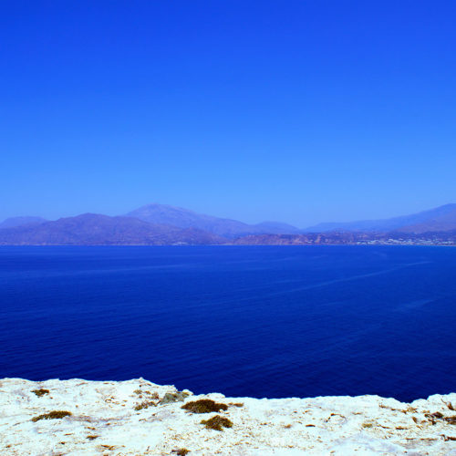 Wide view of a calm blue sea with distant mountains and a rocky shoreline in the foreground