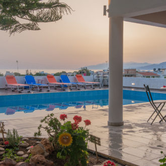 Poolside terrace with colorful loungers, a small table setting, and a sunset view toward the sea and hills
