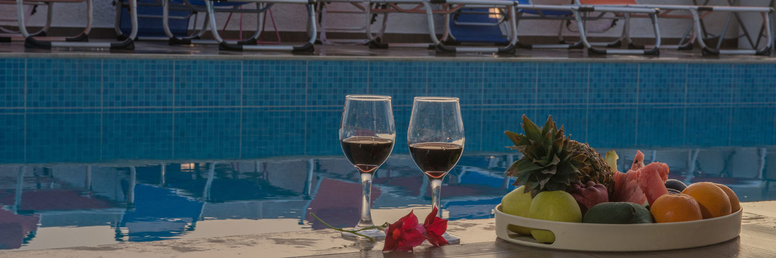 Two wine glasses and a tray of fresh fruit beside a pool with sun loungers at sunset