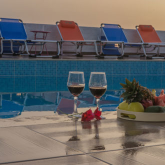 Two wine glasses and a tray of fresh fruit beside a pool with sun loungers at sunset
