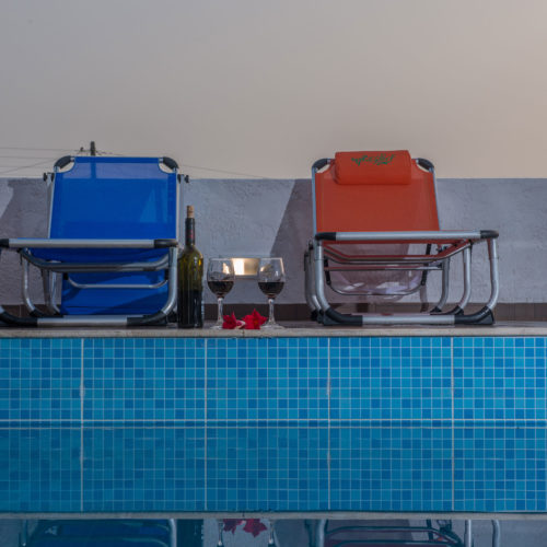 Two lounge chairs sit beside a pool with wine glasses, a bottle, and flowers arranged on the deck
