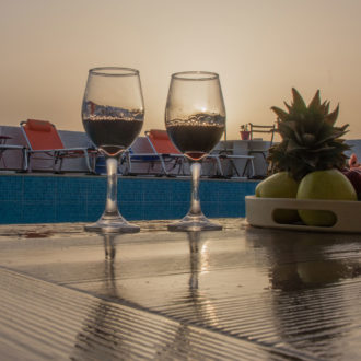 Two wine glasses on a wet table beside the pool at sunset, with lounge chairs and a fruit bowl nearby
