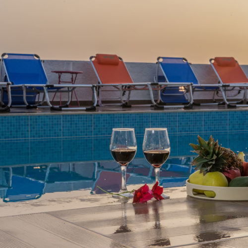 Two wine glasses and a fruit tray beside a pool with sun loungers at sunset