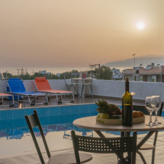 Poolside terrace with sun loungers, a table set with fruit and wine, and a sunset view over the sea
