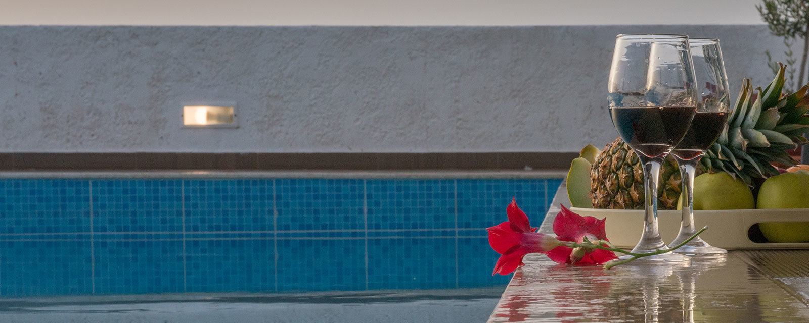 Two glasses of red wine sit beside a tray of tropical fruit and a red flower by a blue swimming pool