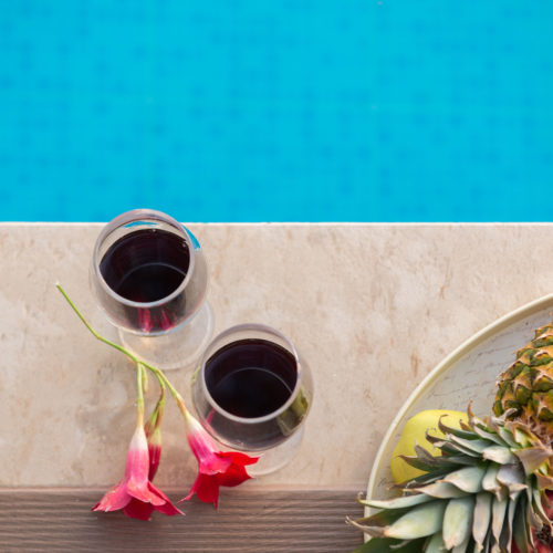 Two glasses of red wine beside tropical fruit and flowers on a poolside table at Houmeli Apartments