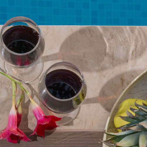 Two glasses of red wine beside tropical fruit and pink flowers on a sunlit table by the pool
