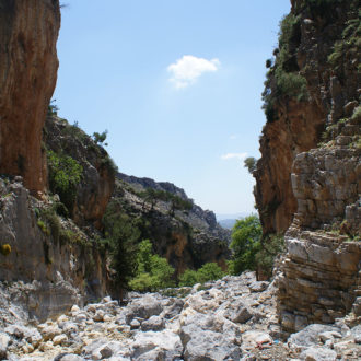 Rocky gorge with steep cliffs, scattered shrubs, and a bright blue sky in South Crete