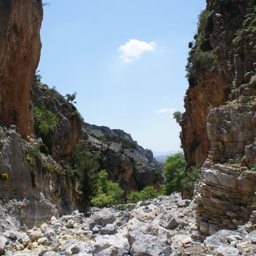 Rocky gorge with steep cliffs, scattered shrubs, and a bright blue sky in South Crete