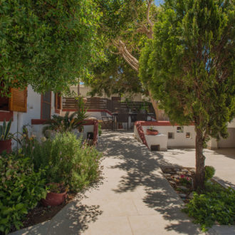 Shaded courtyard at Houmeli Apartments with trees, potted plants, a paved path, and outdoor seating